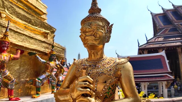 Golden deity statue holding a lotus flower at the Bangkok Grand Palace, surrounded by ornate Thai temple spires under clear blue sky.