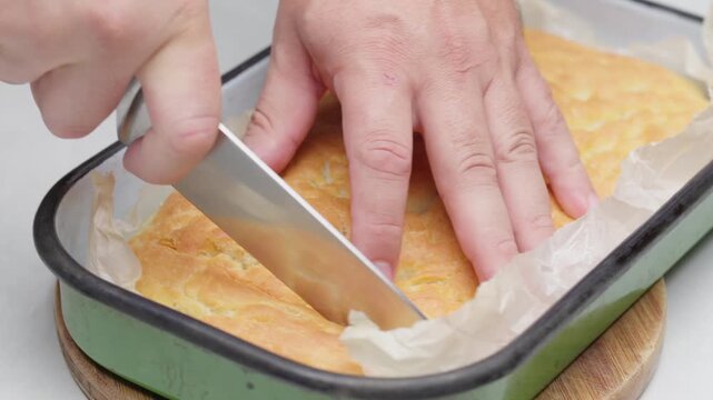 Close-up of hands cutting golden focaccia bread inside a rectangular baking dish