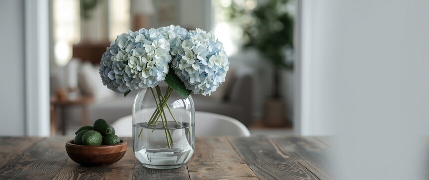 On a dining table, a clear glass vase holding pale blue and white hydrangeas is placed beside a wooden bowl filled with green avocados, with a living room softly out of focus behind