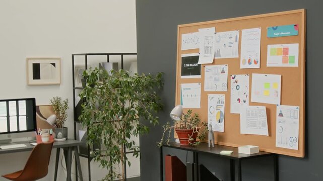 Full no people shot of cork pin board at UX software design company, displaying workflow graphs for project teamwork planning, next to employee desk with computer and verdant green potted plant
