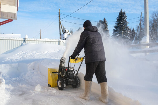 A man clears snow in his backyard using a yellow snow blower during a winter day in a snowy area