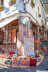 Fototapeta premium Vibrant corner shop in a European old town displaying handmade woven rugs, patterned textiles, magnets, bracelets, and cultural souvenirs outside a charming white stone building with balconies
