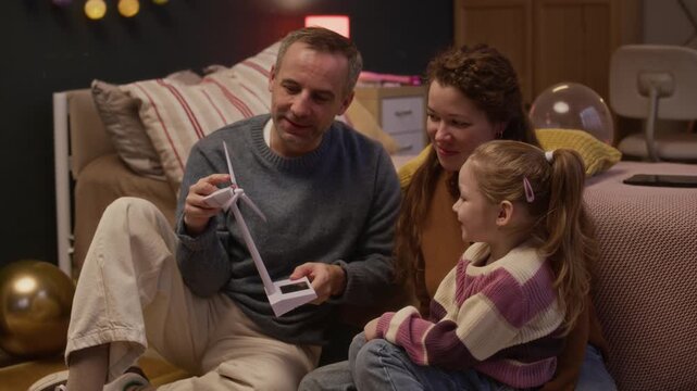 Tilt shot of Caucasian father taking windmill model and showing it to little daughter who sitting near mother