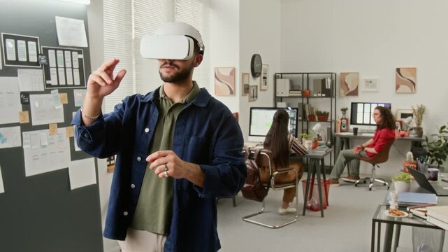 Medium shot of young male user interface designer, wearing AR headset, developing wireframes in metaverse, with drag, scroll, swipe, tap gestures, team colleagues talking by computers in background