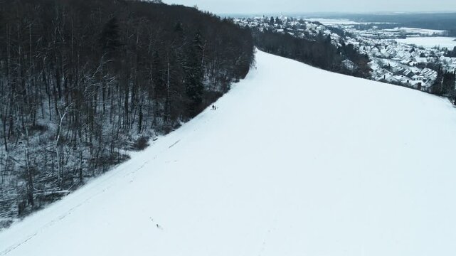Forward drone flight above wide snow-covered fields near Elchingen, with forest edge, distant village and forest, and winter horizon under overcast sky.