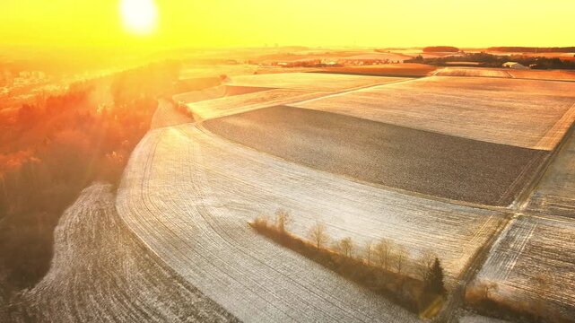 Forward drone flight near Elchingen during golden-hour sunset, gliding over rolling farmland and frosty fields with warm sun flares and long shadows.