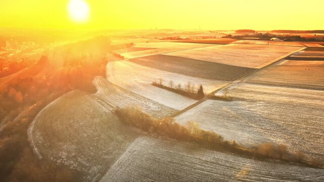 Forward drone flight near Elchingen during golden-hour sunset, gliding over rolling farmland and frosty fields with warm sun flares and long shadows.