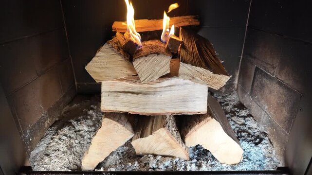 a hand carefully placing a piece of firewood inside an enclosed fireplace. The stacked wooden logs rest on ash-covered ground, highlighting the preparation stage before lighting a fire