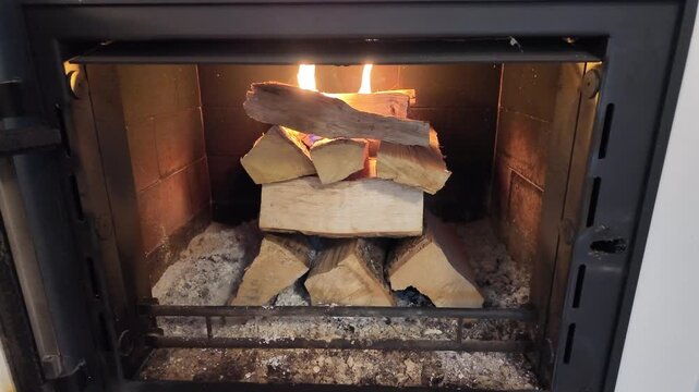 A stacked arrangement of firewood burns inside a closed indoor fireplace, with small flames visible at the top of the wooden logs. Ash and embers cover the base of the firebox