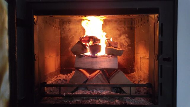 A bright, steady fire burns inside a modern enclosed fireplace, with stacked firewood glowing as tall flames rise in the center. The warm orange light reflects off the fireproof interior,