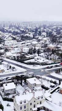 Vertical drone shot over a snow-covered suburb and busy intersection, cars moving through winter haze with light flurries, showing residential rooftops and commute routes.