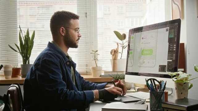 Medium side shot of focused young biracial male user experience designer, wearing denim jacket, drawing layouts for mobile app using graphic tablet and stylus, displayed on computer monitor