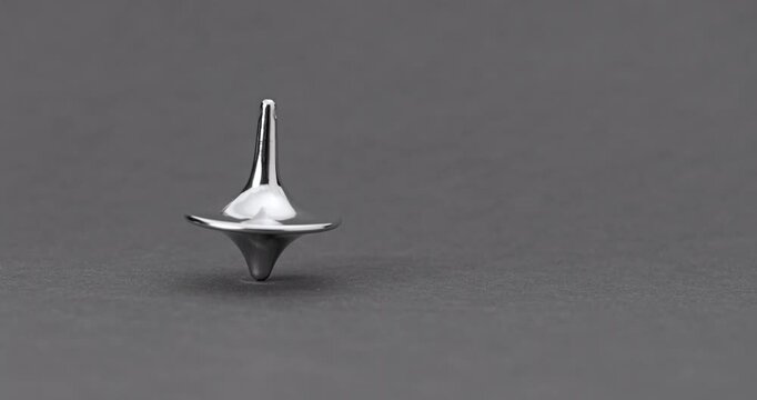 Metallic Spinning Top Falling Over. Linear footage of a shiny chrome top in steady rotation, progressively wobbling with increasing tilt until it fully topples on a matte gray surface.