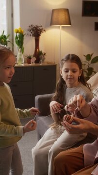 Vertical shot of little Caucasian girls helping their grandmother to wrap thread around dyed Easter egg, chatting and having fun while preparing for spring holiday together at home