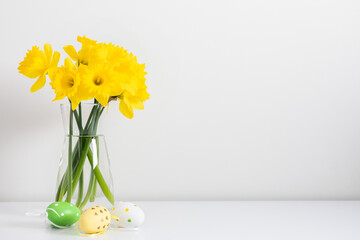 Easter composition. A bouquet of yellow daffodils in a glass vase and decorative Easter eggs on a white background. Spring Easter background for a card or banner