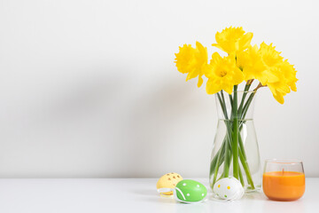 Easter background. A bouquet of yellow daffodils in a clear vase, complemented by three decorative Easter eggs and an orange candle on a white table. Bright Easter spring composition.