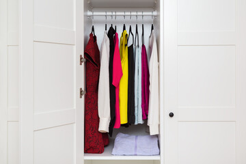 Colorful different female clothes hanging on black hangers inside opened white wooden wardrobe at home room. Closeup. Front view.