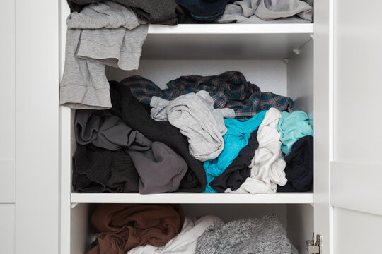 Cluttered different colorful male clothes on shelves in white opened wooden wardrobe at home room. Messy casual apparel. Closeup. Front view.