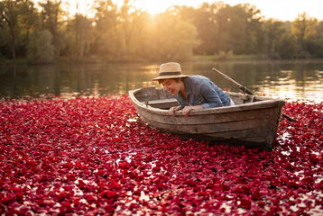 Excited young woman in a wooden boat looking at a lake surface covered with thousands of red rose petals during a romantic golden hour sunset for Valentine's Day