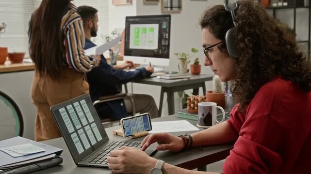 Medium side shot of curly young Caucasian male UI designer viewing app wireframes on tablet and smartphone, drinking coffee, team colleagues discussing project issues in background