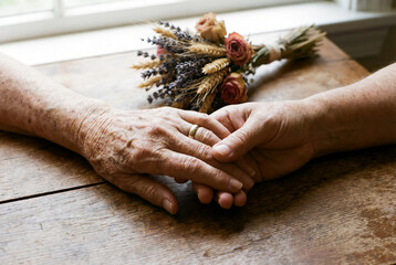 Close up of elderly couple hands with gold wedding ring holding each other on wooden table with dried flowers symbolizing eternal love and care for Valentine's Day