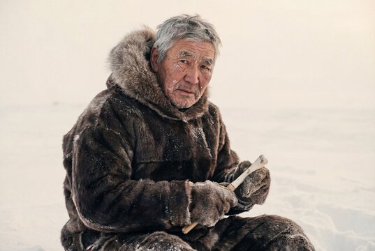 Elderly inuit hunter wearing traditional fur clothing sitting in snow holding a bone tool outdoors