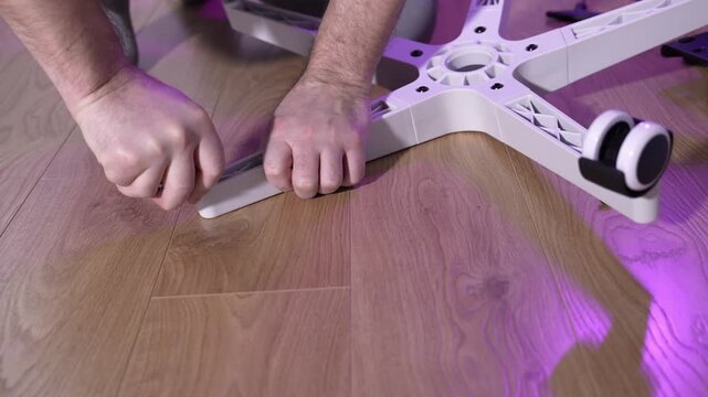 Hands attaching caster wheels to white plastic office chair base on wooden floor. Furniture assembly process, office chair setup, indoor DIY work, close-up, no face.