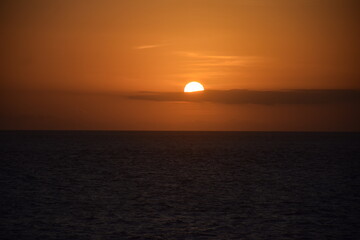 Fototapeta premium The sun appears round, surrounded by long, black clouds, and the orange sky is setting as the sun sets on the western horizon.