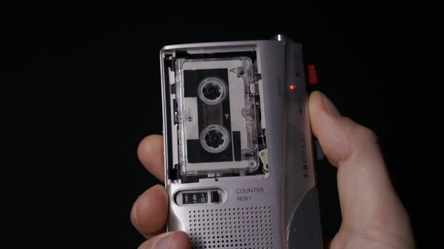 A hand holds a silver microcassette recorder against a black background and presses play to start the tape. Concept of retro technology, investigative journalism, and analog audio evidence.