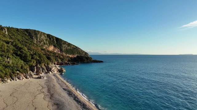 Dhermi, Albania - April 08, 2025: Aerial view of a serene beach with gentle waves lapping against the shore, surrounded by lush green hills under a clear blue sky