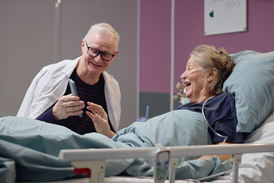 Senior Caucasian man showing smartphone to senior Caucasian woman lying in hospital bed, both smiling and interacting, medical setting indicating vascular center patient care scenario