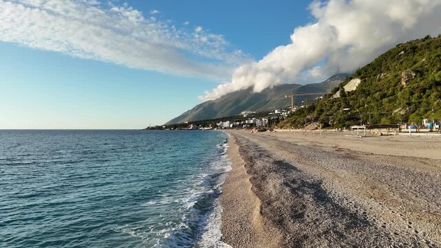 Dhermi, Albania - April 08, 2025: Scenic coastal view of a pebbled beach with clear blue water, mountains, and clouds under a bright sky in a serene seaside location