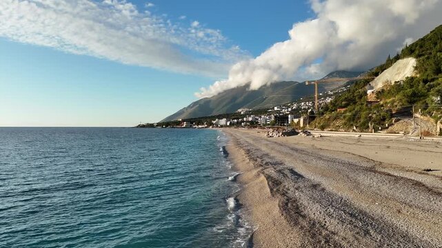 Dhermi, Albania - April 08, 2025: Scenic coastal view of a beach with gentle waves lapping at the shore, mountains and buildings visible under a clear blue sky