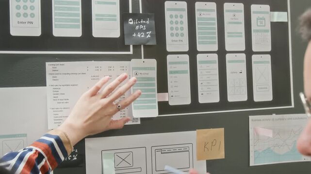 Rear close-up of female project manager pointing to options on wireframe layout, hanging on wall display board in startup office, while brainstorming ideas for developing mobile app