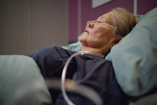 Senior Asian woman lying in hospital bed receiving oxygen therapy through nasal cannula, resting with eyes closed in vascular center, medical tubing visible near patient