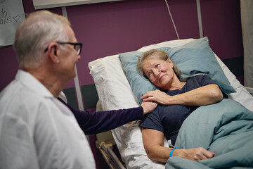 Obraz premium Senior Caucasian woman lying in hospital bed receiving comfort from senior Caucasian man holding her hand in vascular center patient room