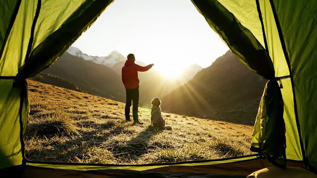A captivating view from inside a camping tent reveals a man and his loyal dog silhouetted against a breathtaking golden sunrise over majestic snow-capped mountains and a vast grassy valley. The man is