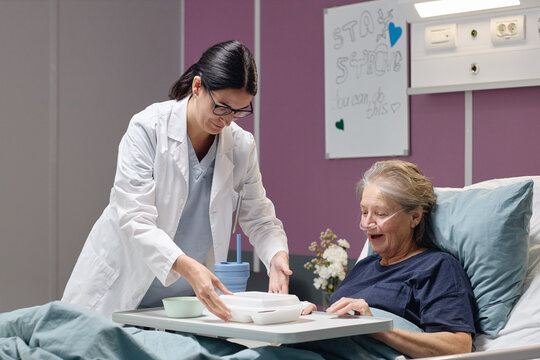 Caucasian female doctor serving meal to senior Caucasian woman lying in hospital bed, both smiling, medical equipment and motivational sign visible in vascular center patient room