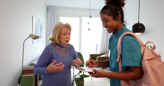 Professional female home nurse visiting an elderly woman at her house, asking questions about her health and taking notes on a clipboard during a routine medical examination and consultation