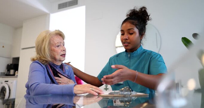Empathetic young female caregiver providing comfort and emotional support to a senior woman, holding her hand during a home visit and offering reassuring words while sitting at a table together