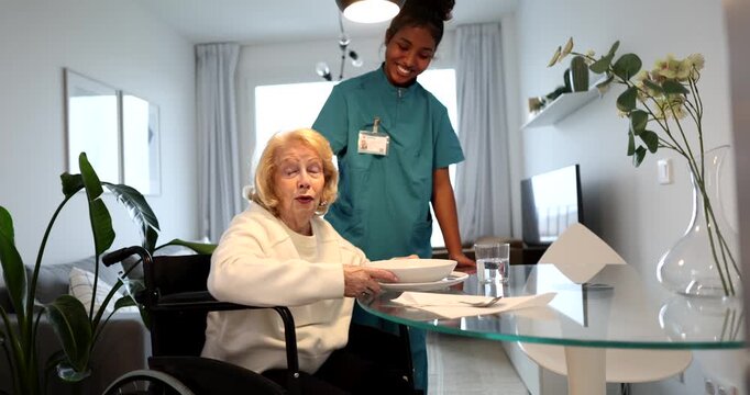Young. Smiling female caregiver assisting an elderly woman in a wheelchair. Bringing her a plate of food at the dining table in a modern apartment. Illustrating home care assistance and companionship
