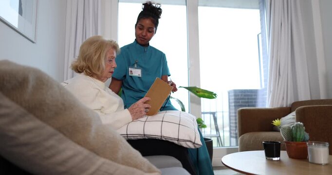Compassionate african american nurse reading a book aloud to a senior woman, providing comfort and companionship in a cozy nursing home living room during a home care visit