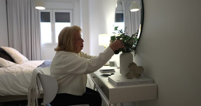 Senior woman sitting at a white vanity table in a cozy bedroom, carefully arranging a bouquet of flowers in a vase, adding a touch of nature and elegance to her personal space