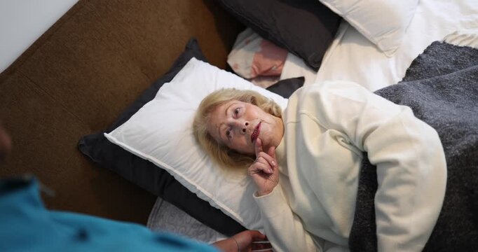 Elderly woman lying in bed and talking to her caregiver during a home visit. The medical professional, wearing blue scrubs, sits by her bedside, providing comfort and support to the senior patient
