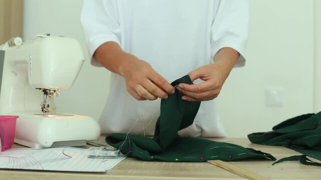 A person hand-sews dark green fabric with a needle and thread, preparing seams beside a sewing machine, pins, and pattern paper on a wooden table.