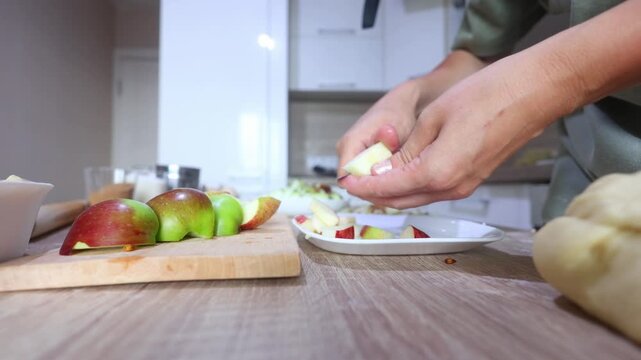 A person slices a red apple into thin wedges on a wooden board, precise, rhythmic cuts in a bright kitchen, dough and ingredients nearby.