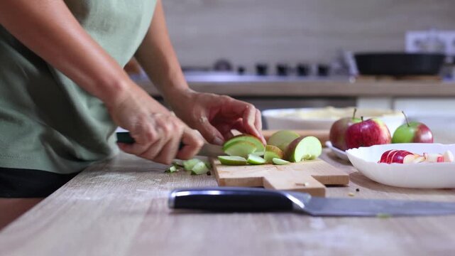 A person slices a red apple into thin wedges on a wooden board, precise, rhythmic cuts in a bright kitchen, dough and ingredients nearby.