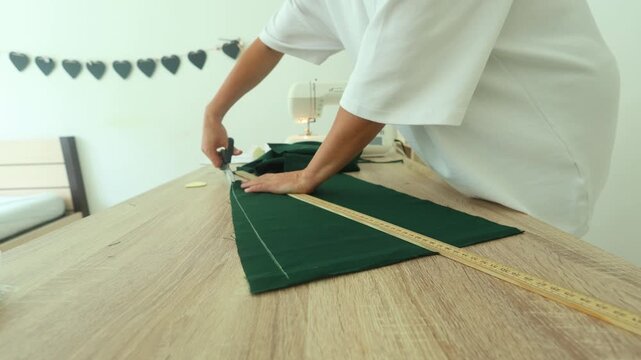 A person measures and cuts deep green fabric with a ruler and scissors on a wooden table, focused, precise sewing prep in a bright