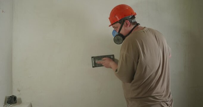 A professional builder in a hard hat and respirator sands a freshly plastered wall with a hand sander. A man sands a plastered wall to prepare the surface for painting during a home renovation.