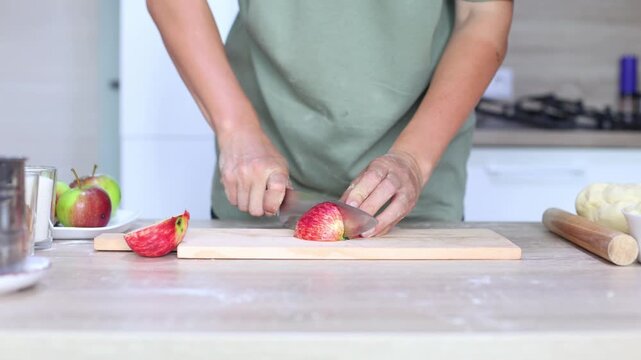 A person slices a red apple into thin wedges on a wooden board, precise, rhythmic cuts in a bright kitchen, dough and ingredients nearby.